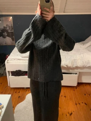 Mörkgrått stickat set med kjol - Säljer ett mörkgrått stickat set med långärmad tröja och matchande lång kjol. Setet har en ribbstickad struktur och tröjan har vida ärmar. Perfekt för dig som gillar en avslappnad och trendig look.