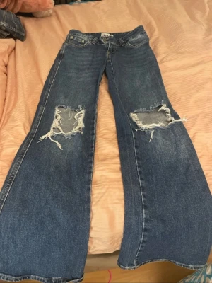 Blå bootcut jeans med slitningar - Säljer ett par blå bootcut jeans med stora slitningar över knäna. Jeansen har en klassisk femficksmodell och är tillverkade i ett robust denimtyg. Perfekta för dig som gillar en avslappnad och trendig stil.