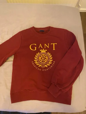 Röd sweatshirt från GANT - Snygg röd sweatshirt från GANT med stort gult tryck och logga på bröstet. Tröjan har rund halsringning, ribbade muddar vid ärmslut och nederkant samt en klassisk relaxed fit. Perfekt för en sportig och trendig look. Bara att skriva vid frågor och priset går såklart att diskutera!