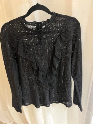 Svart spetsblus med volangdetaljer - Svart blus i spets med långa ärmar och volangdetaljer framtill