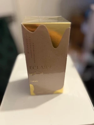 Lattafa Eclaire Eau de Parfum - Lattafa Eclaire Eau de Parfum kommer i en lyxig beige och guld kartong med elegant, glansig finish och gulddetaljer. Förpackningen har en modern design med mjuka former och text i guld. Perfekt för dig som gillar exklusiva dofter och snygg design.