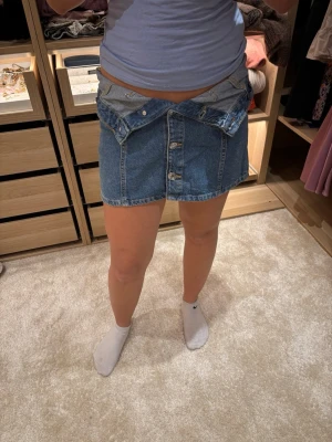 Blå jeanskjol med knappar framtill - Säljer en klassisk blå jeanskjol med knappar hela vägen framtill och två stora fickor. Kjolen har en rak passform och är i ett kraftigt denimtyg som ger en cool och avslappnad look. Perfekt till t-shirt eller hoodie. Kan ha den på olika sätt!