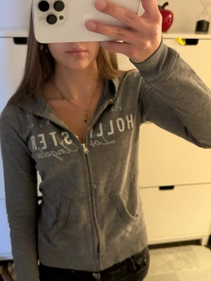  Hollister zip up hoodie  - Grå huvtröja med dragkedja från Hollister. Klassisk modell med huva, fickor framtill och vit textlogga på bröstet. Mjuk bomullsblandning och ribbade muddar vid ärmslut och nederkant. Perfekt för en chill och avslappnad stil.