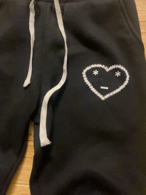 Svarta mjukisbyxor Carsicko - Svarta mjukisbyxor med vit snörning i midjan och ett broderat hjärta med ansikte på ena benet. Byxorna har en relaxed passform och är perfekta för chill dagar. Tillverkade i mjukt material för extra komfort.