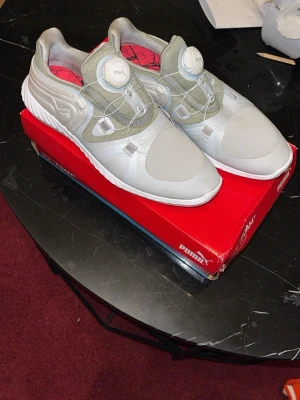 Puma sneakers i ljusgrått och vitt - Snygga Puma sneakers i ljusgrått och vitt med unikt snörningssystem och detaljer i olivgrönt. Insidan har ett rött mönster och ovandelen är i syntetmaterial. Perfekta för dig som vill ha något sportigt och stilrent.