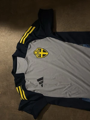 Svensk tränings t-shirt Adidas S - Snygg tränings t-shirt från Adidas i grått och mörkblått med gula detaljer och Sveriges landslagsemblem på bröstet. Tillverkad i funktionsmaterial som andas, perfekt för träning eller sport. Slim fit-modell med klassiska Adidas-ränder på axlarna.
