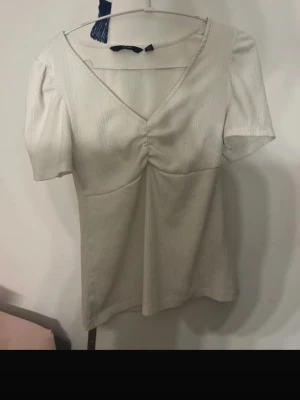 Vit v-ringad topp från Vero Moda - Snygg vit topp från Vero Moda med v-ringning och lätt puffade korta ärmar. Toppen har en rynkad detalj framtill och är tillverkad i ett mjukt, strukturerat material. Perfekt att styla med jeans eller kjol för en trendig look.