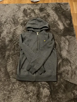 Mörkgrå zip hoodie från Lyle & Scott - Mörkgrå hoodie med dragkedja från Lyle & Scott. Klassisk modell med huva, två fickor framtill och gul logga på bröstet. Tillverkad i mjukt bomullsmaterial som är skönt mot huden. Perfekt för en avslappnad och stilren look.