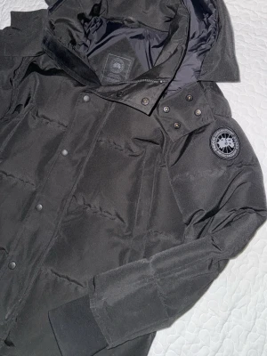 Svart dunjacka från Canada Goose  - Säljer en svart dunjacka från Canada Goose i storlek XS. Jackan har huva, ribbade muddar och tryckknappar framtill. Klassisk patch på ärmen och quiltad design som håller dig varm under kalla dagar. Perfekt för dig som vill ha en stilren och funktionell vinterjacka. Jackan passar till passform S och M bättre 