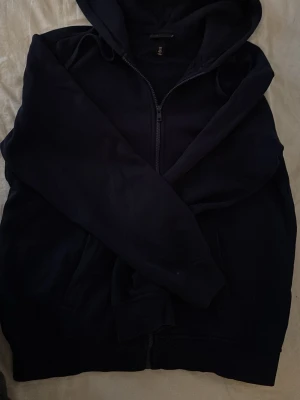 Svart hoodie med dragkedja från H&M - Mörkblå hoodie från H&M i relaxed fit med dragkedja framtill och justerbar huva. Tillverkad i mjukt bomullsmaterial och har fickor på sidorna. Perfekt för en avslappnad och stilren look.