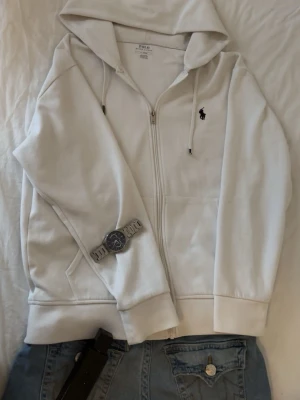 Vit hoodie från Polo Ralph Lauren - Säljer en stilren vit hoodie från Polo Ralph Lauren med dragkedja framtill och klassisk huva. Hoodien har det ikoniska svarta Polo-logotypen broderad på bröstet och praktiska fickor. Tillverkad i mjukt bomullsmaterial för maximal komfort.