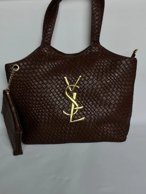 Brun YSL handväska i skinn - Snygg brun handväska från YSL i flätat skinn med stort guldfärgat YSL-emblem framtill. Väskan har två handtag och medföljande liten clutch med guldkedja. Perfekt för dig som vill ha en lyxig och rymlig väska med exklusiv känsla.