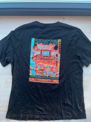 Svart t-shirt Urban Threads strl L med ryggtryck - Säljer en svart t-shirt från Urban Threads med stort färgglatt Chinatown-tryck på ryggen och texten 'CHINATOWN NEW YORK CITY' framtill. Skön passform och klassisk rund hals. Perfekt för dig som gillar streetwear och grafiska prints.