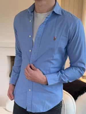 Blå skjorta från Ralph Lauren - Klassisk ljusblå skjorta från Ralph Lauren med broderad logga på bröstet. Skjortan har knappar framtill och långa ärmar med manschett. Perfekt för en stilren och avslappnad look.