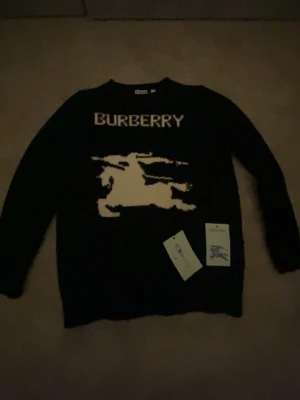 Svart stickad tröja från Burberry - Perfekt för vintern skön Stickad svart tröja från Burberry med stort vitt Burberry-tryck och riddarlogga på bröstet. Tröjan har rund halsringning och långa ärmar. Perfekt för dig som gillar klassiska designerplagg med en modern twist. Pris kan diskuteras 🏆 