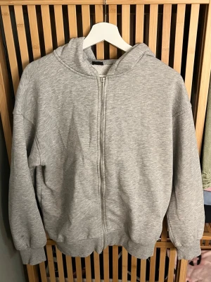 Grå zip-hoodie med huva - En klassisk grå hoodie med dragkedja och huva. Modellen har ribbade muddar vid ärmslut och nederkant samt en mjuk, bekväm känsla. Perfekt för en avslappnad stil och enkel att matcha med andra plagg.