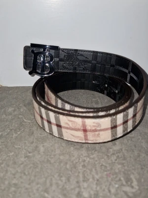 Burberry Bälte (Reversible) - Vändbar 2 sidig Burberry bälte med klassiskt rutmönster i beige och svart. Bältet har en svart och silvrig metallspänne med Burberry loggan på. Perfekt accessoar för att lyfta din outfit med en lyxig touch. UTAN BOX!