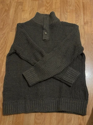 Mörkgrön stickad tröja med krage - Stickad tröja i mörkgrönt med hög ribbad krage och knappdetalj framtill. Grovstickad struktur och breda muddar vid ärmslut och nederkant. Perfekt för kyliga dagar och enkel att matcha med jeans.