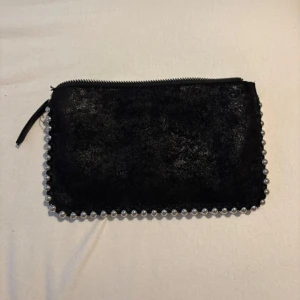 Svart kuvertväska med silverpärlor - Snygg svart kuvertväska i mocka-look med dragkedja och coola silverfärgade pärlor längs kanterna. Perfekt storlek för mobilen och det viktigaste. Stilren och edgy design som sticker ut. Väskan har något form av smuts inuti men annars är den fräsch. 
