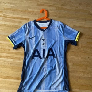 Tottenham Hotspur blå fotbollströja Nike - Snygg Tottenham Hotspur fotbollströja från Nike i olika blå nyanser med vertikala ränder. Kortärmad modell med mörkblå krage och detaljer i gult vid ärmslut. Stor AIA-logga på bröstet och klubbens emblem. Tillverkad i lätt och ventilerande polyester.