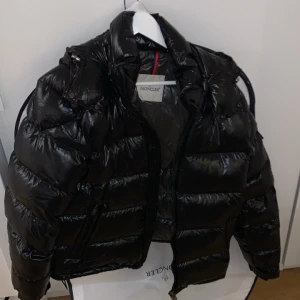 Moncler jacka - Svart pufferjacka från Moncler med glansig finish och huva. Jackan har Moncler-logga på ärmen, dragkedja framtill och flera fickor. Perfekt för kalla dagar och har en riktigt snygg streetstil. Kommer med original Moncler klädpåse. Storlek 3. Nyskick