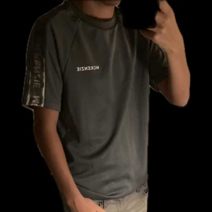 Grå t-shirt från McKenzie - Snygg grå t-shirt från McKenzie med korta ärmar och logga på bröstet. Den har en stilren design med en randig detalj längs ärmarna. Perfekt för en avslappnad stil.