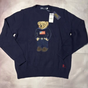 Marinblå stickad tröja Polo Bear - Snygg marinblå stickad tröja från Polo Ralph Lauren med ikoniska Polo Bear broderad framtill. Rund hals, ribbade muddar och RL-broderi i rött vid nederkanten. Perfekt för dig som vill ha en klassisk men lekfull look.