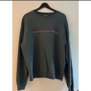 Mörkgrön sweatshirt från Polar Skate Co - Sweatshirt i mörkgrönt från Polar Skateboard Company, med broderade röda figurer och vit text på bröstet. Tröjan har rund hals, ribbade muddar och lång ärm. Perfekt för dig som gillar streetwear och skate.