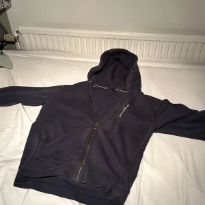 Mörkblå hoodie från Sail Racing - Mörkblå hoodie från Sail Racing i storlek M. Tröjan har huva, hel dragkedja och mjukt fleecefoder på insidan. Långärmad modell med logotyp broderad på bröstet. Perfekt för kyliga dagar och chill stunder.