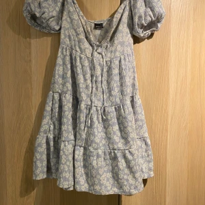 Blommig ljusblå kortklänning Gina Tricot - Supersöt ljusblå kortklänning från Gina Tricot med vitt blommigt mönster. Klänningen har puffiga kortärmar, v-ringning och volanger i kjolen. Tillverkad i mjuk bomull med smockad struktur och knyt framtill. Perfekt för sommarens alla tillfällen!