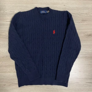 Mörkblå kabelstickad tröja Polo Ralph Lauren - Snygg mörkblå kabelstickad tröja från Polo Ralph Lauren i storlek M. Tröjan har ett klassiskt rött broderat Polo-logo på bröstet, rund hals och ribbade muddar. Tillverkad i 100% bomull för skön och mjuk känsla. Perfekt för dig som gillar stilrena och tidlösa plagg.