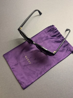 Gucci solglasögon - Snygga svarta solglasögon från Gucci, modell CG0636SK. Cat-eye form med guldfärgade detaljer och GG-logga på skalmarna. Kommer med lila Gucci-fodral i satin. Perfekt accessoar för att lyfta din look och skydda ögonen mot solen.