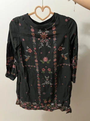 Svart blus med broderade blommor - Svart blus med färgglada broderade blommor och fåglar framtill och på ärmarna. Blusen har trekvartsärm med lätt puff och elastisk kant. Rund halsringning och detaljerad brodyr längs nederkant och ärmslut. Perfekt för dig som gillar boho vibes och unika detaljer.