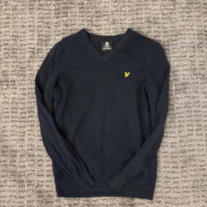 Marinblå v-ringad tröja Lyle & Scott - Snygg marinblå v-ringad tröja från Lyle & Scott med gul broderad logga på bröstet. Tröjan har långa ärmar och ribbade muddar vid ärmslut och nederkant. Perfekt för dig som gillar stilrena och klassiska plagg med en modern touch.