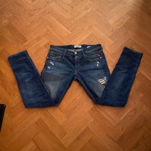 Blå Replay jeans med slitningar - Snygga blå jeans från Replay med coola slitningar och klassisk femficksdesign. Jeansen har en normal passform och raka ben, med detaljer som ljusa tvättade partier och Replay-logga på bakfickan. Perfekta för dig som gillar en avslappnad streetstil.
