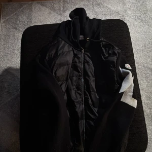 Svart dunjacka från Moncler med huva - Svart dunjacka från Moncler med huva och dragkedja framtill. Jackan har ett coolt vitt zigzag-mönster på ena ärmen och Moncler-logga på axeln. Materialet är en mix av dun och polyester, perfekt för kalla dagar. Snygg och modern look med quiltade detaljer.