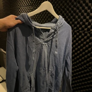 Blå hoodie från ralph lauren med knappar - Blå hoodie från Ralph lauren med huva och snörning, samt knappslå vid halsen. Tröjan har en känguruficka framtill och ett diskret broderat emblem på bröstet. Tillverkad i mjuk bomull, perfekt för chill dagar. Långärmad och loose passform.