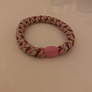 Färgglatt flätat armband med rosa detalj - Säljer ett coolt flätat armband i beige, rosa och glittriga trådar. Armbandet har en rund form och en rosa plastdetalj med text. Perfekt accessoar för att liva upp din outfit!