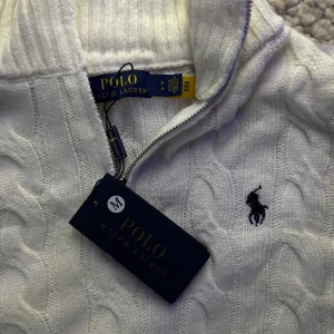 Vit stickad tröja Polo Ralph Lauren - Säljer en vit stickad tröja från Polo Ralph Lauren med klassiskt kabelstickat mönster och half zip vid halsen. Tröjan har långärmad design och den ikoniska broderade loggan på bröstet. Perfekt för dig som gillar stilren och tidlös look.