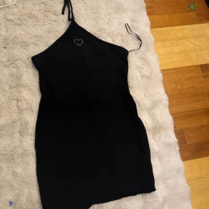 Svart kort klänning med hjärtdetalj - Svart kort klänning med smala axelband och asymmetrisk halsringning. På framsidan finns en liten hjärtdetalj i vitt. Klänningen är tight och har en enkel, stilren design som passar dig som gillar minimalistiskt mode.