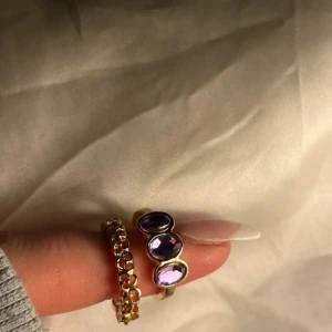 Två guldiga ringar med lila stenar - Säljer två snygga ringar i guldfärgad metall. Den ena har ett coolt kedjemönster runt hela ringen, medan den andra har tre ovala lila stenar som verkligen poppar. Perfekt för dig som gillar att mixa och matcha smycken med färg och detaljer.