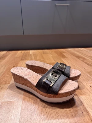 Michael Kors sandaler med korkkilklack - Snygga sandaler från Michael Kors med breda mörkbruna remmar i skinn och guldfärgade MK-detaljer framtill. Sulan är i kork med vit och brun plattform, vilket ger extra höjd och en cool look. Perfekta för sommarens alla tillfällen.