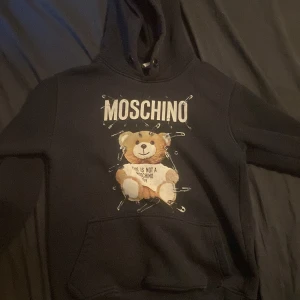 Svart Moschino hoodie med nalletryck. - Svart hoodie från Moschino med stort tryck av en gullig nalle och säkerhetsnålar på framsidan. Tröjan har huva och en klassisk känguruficka. Perfekt för dig som gillar statement-plagg och coola designer. Materialet är mjukt och skönt, troligen bomull.