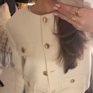 Vit kavajjacka från Zara med guldknappar - Säljer en stilren vit kavajjacka från Zara med rund halsringning och snygga guldknappar framtill. Jackan har fyra fickor med lock och är tillverkad i ett strukturerat tyg som ger en exklusiv känsla. Perfekt för dig som gillar klassisk och chic stil.