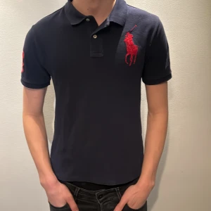 Mörkblå pikétröja från Ralph Lauren - Snygg mörkblå pikétröja från Ralph Lauren med röd broderad logga på bröstet och en stor röd siffra 3 på ärmen. Klassisk krage med knappar och korta ärmar. Perfekt för en stilren och sportig look. Modellen är 186 cm och väger 70kg 