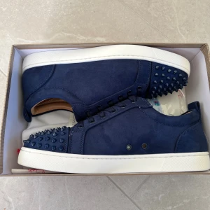 Mörkblå sneakers med nitar från Louboutin - Säljer ett par mörkblå sneakers från Louboutin i mocka med coola ton-i-ton nitar på tån. Skorna har snörning, vit platt sula och den klassiska röda undersidan. Insidan är beige och de har en rund tå. Perfekt för dig som gillar edgy detaljer och lyxig design. Finns i båda 42 och 43