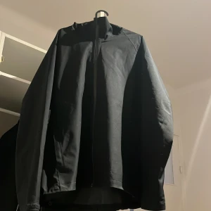 Svart vindjacka från Uniqlo XL - Svart vindjacka från Uniqlo i storlek XL. Jackan har hög krage, dragkedja framtill och är tillverkad i ett vattenavvisande och vindtätt syntetmaterial. Enkel och stilren design med långärmade ärmar, perfekt för blåsiga dagar.