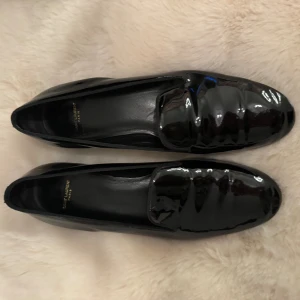 Svarta lackloafers från Saint Laurent - Snygga svarta loafers från Saint Laurent i glansigt lackskinn. Klassisk rund tå och låg klack ger en stilren siluett. Perfekta för dig som gillar exklusiva och tidlösa skor med lyxig finish. Tillverkade i Italien.