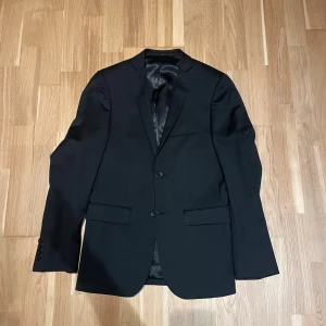 Svart klassisk kavaj med knappar från dressman  - Snygg svart kavaj med två knappar framtill och klassisk krage. Fickor med lock och slät design ger en stilren look. Perfekt för dig som vill ha en enkel och tidlös kavaj till garderoben.