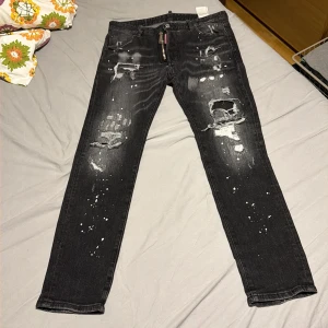 Svarta Dsquared2 jeans med slitningar storlek 52 - Säljer ett par svarta jeans från Dsquared2 med coola slitningar, hål och vit färgstänk över hela byxorna. Jeansen har en rak passform och klassisk femficksdesign. Materialet är jeans och de har en modern, edgy look. Storlek 52